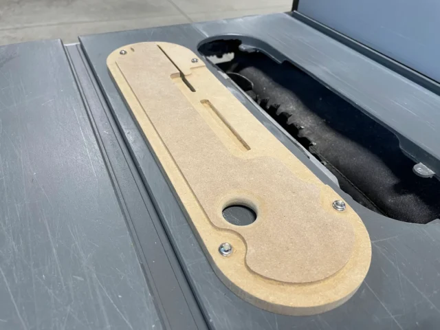 Tan MDF zero clearance insert for Dewalt table saw, displayed in the saw's slot next to black dust collection tube, showing mounting holes and curved edges