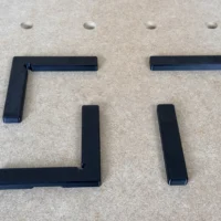 Four black 3D-printed fence pieces for 20mm workbench grid: two L-shaped corner fences, two straight fence rails, shown disassembled on pegboard.