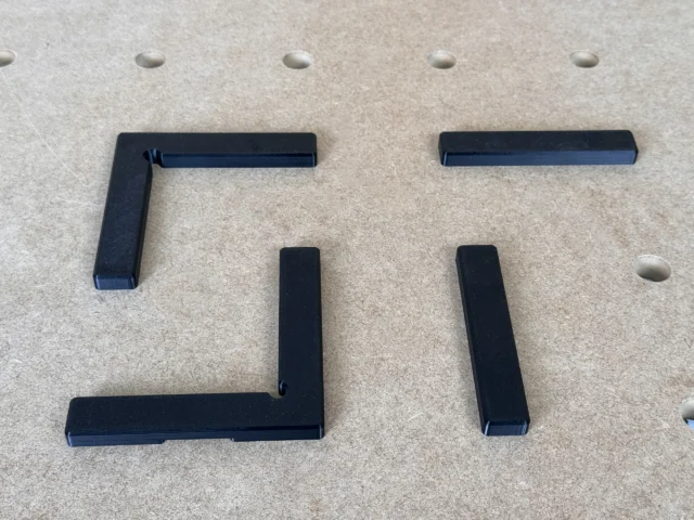 Four black 3D-printed fence pieces for 20mm workbench grid: two L-shaped corner fences, two straight fence rails, shown disassembled on pegboard.
