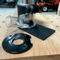 Makita trim router extended base plate with black curved mounting plate and clear dust collection cup shown separately on workbench