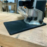Black extended base plate attached to Makita trim router with clear dust collection shroud, shown from front angle on wooden surface