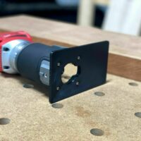 Black extended base plate attached to a Milwaukee trim router with red housing, shown on a work surface with dowel holes