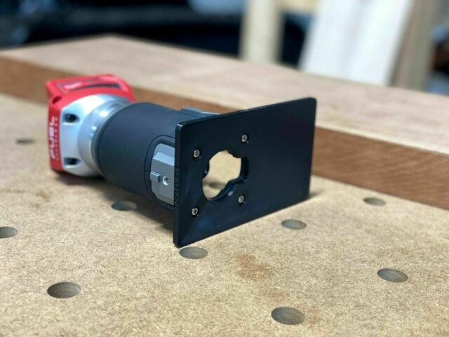 Black extended base plate attached to a Milwaukee trim router with red housing, shown on a work surface with dowel holes