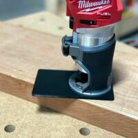 Black extended base plate mounted on Milwaukee Fuel M18 trim router, positioned on wooden surface with alignment holes visible