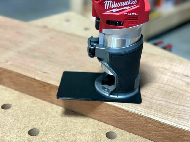 Black extended base plate mounted on Milwaukee Fuel M18 trim router, positioned on wooden surface with alignment holes visible