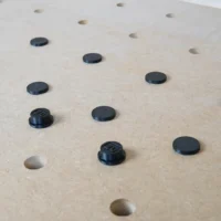 Black button protectors installed in 20mm bench dog holes on a light wood workbench surface, with some holes empty and two protectors showing threaded bases