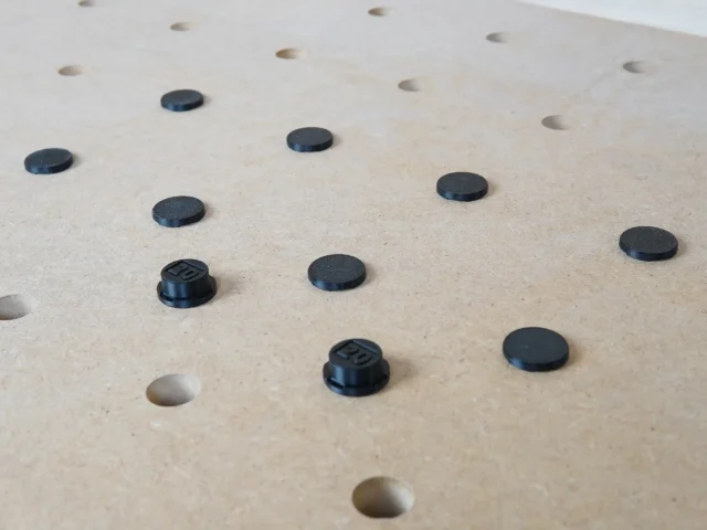 Black button protectors installed in 20mm bench dog holes on a light wood workbench surface, with some holes empty and two protectors showing threaded bases