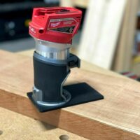 Milwaukee Fuel compact router with red motor head mounted on black rectangular extended base plate, positioned on wooden surface