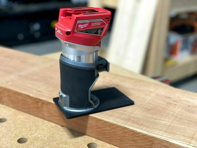 Milwaukee Fuel compact router with red motor head mounted on black rectangular extended base plate, positioned on wooden surface