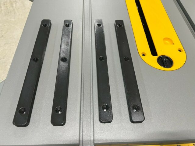 Four black miter bars lined up on a DeWalt table saw surface, each with three countersink holes for mounting