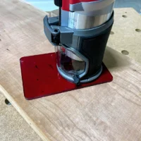Red acrylic base plate attached to Milwaukee trim router, mounted on wooden workbench with holes visible