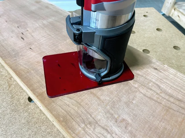 Red acrylic base plate attached to Milwaukee trim router, mounted on wooden workbench with holes visible
