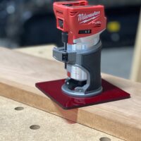 Red acrylic base plate attached to Milwaukee Fuel trim router, mounted on wood with dowel holes visible