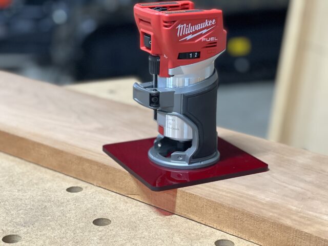 Red acrylic base plate attached to Milwaukee Fuel trim router, mounted on wood with dowel holes visible
