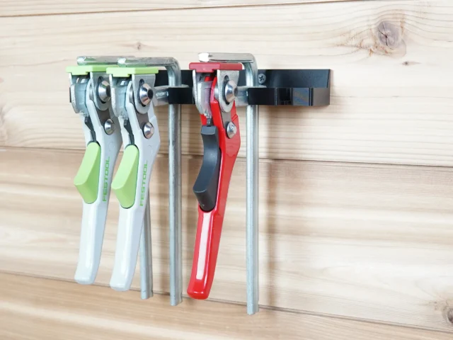 Quick clamp wall mount rack holding four Festool clamps with green and white handles and one red Bessey clamp, mounted on light wood wall