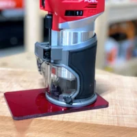 Red acrylic base plate mounted on a Milwaukee M18 trim router, shown from the side on a wooden surface