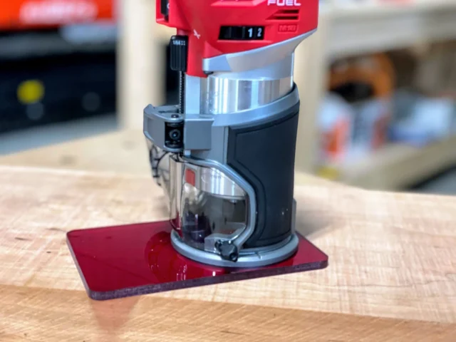 Red acrylic base plate mounted on a Milwaukee M18 trim router, shown from the side on a wooden surface