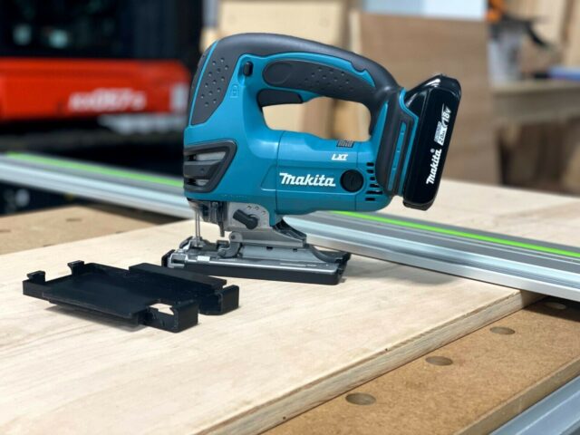 Black snap-on adapter plate detached from blue Makita jigsaw, positioned on wooden surface next to aluminum track rail