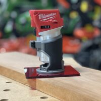 Red acrylic base plate attached to a Milwaukee Fuel trim router, positioned on a wooden workbench with pocket holes visible