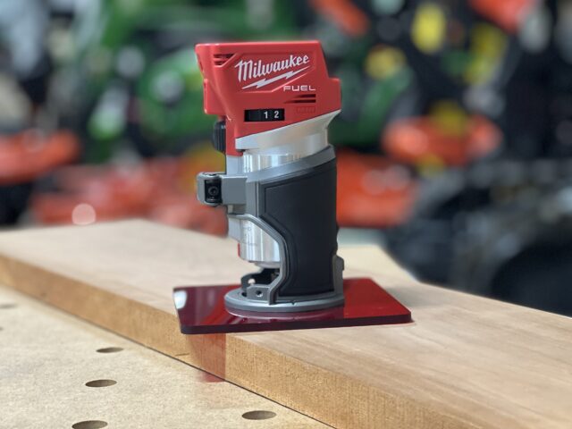 Red acrylic base plate attached to a Milwaukee Fuel trim router, positioned on a wooden workbench with pocket holes visible