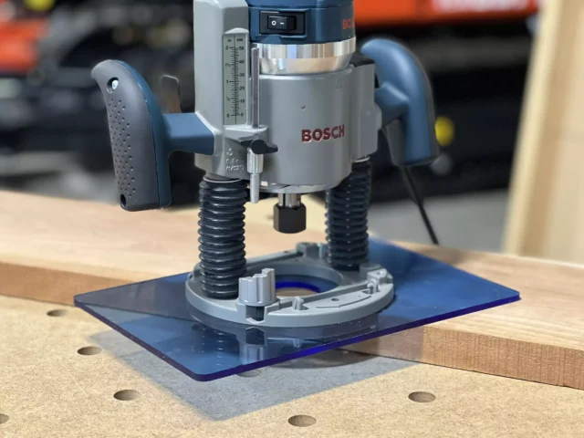 Blue transparent acrylic base plate attached under a Bosch 1617 plunge router on a wooden workbench with guide holes