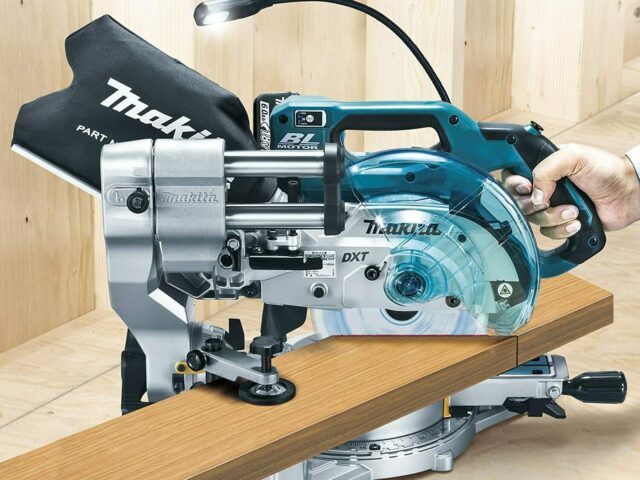 Makita LS610DZ miter saw held in hand while cutting a wooden board, showing the compact blue and silver design with dust collection bag and LED work light