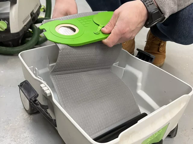 Bright green reusable filter bag held above a white Festool dust extractor container, showing the bag's curved shape and central mounting port