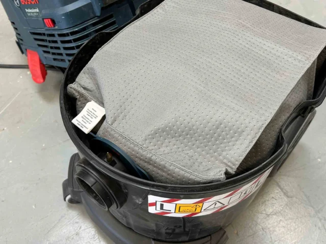 Gray reusable dust bag installed in black Bosch VAC090 9 gallon dust extractor, shown from above with Bosch power tool visible in background