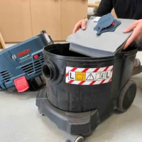 Macam reusable dust bag held above a Bosch VAC090 9-gallon dust extractor, showing gray polyester fleece material and red inner layer