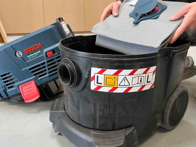 Macam reusable dust bag held above a Bosch VAC090 9-gallon dust extractor, showing gray polyester fleece material and red inner layer