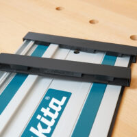 Two black Makita guide rail protective caps fitted on an aluminum rail on a wooden workbench, angled view with ToolCurve markings.