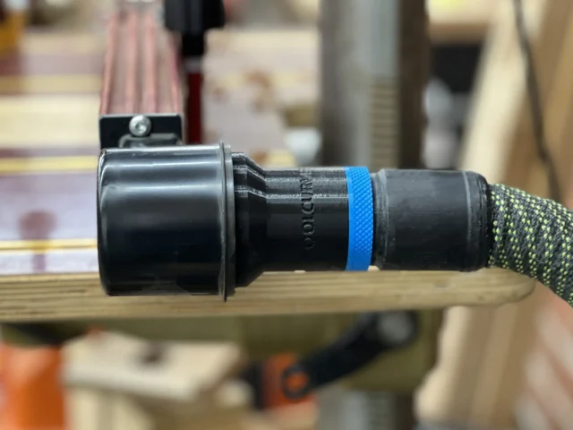 Hose adapter for Woodpeckers DP-PRO fence attached to drill press dust port, showing cylindrical black plastic connector with blue band and braided hose