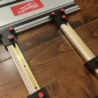 Black parallel guide brackets with red knurled screws mounted on a Milwaukee track saw rail, holding two Incra T-track scales
