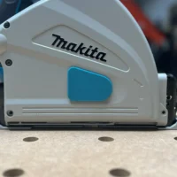 White and blue dust cover attached to the side of a Makita track saw, showing snap-on housing over the arbor area
