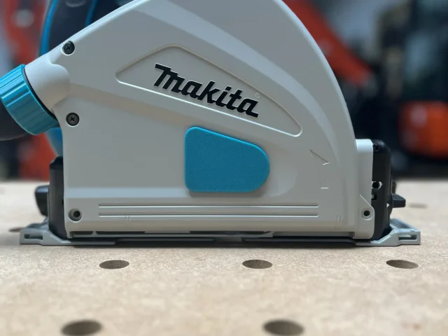 White and blue dust cover attached to the side of a Makita track saw, showing snap-on housing over the arbor area