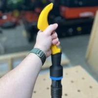 Yellow ProScrapper blade with black hose adapter and blue band attached, held in hand above pegboard workbench