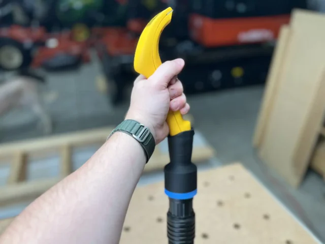 Yellow ProScrapper blade with black hose adapter and blue band attached, held in hand above pegboard workbench