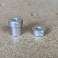 Two aluminum bench dogs side by side, one taller and one shorter, each with threaded top holes and grooved shoulders, on a concrete surface