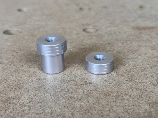 Two aluminum bench dogs side by side, one taller and one shorter, each with threaded top holes and grooved shoulders, on a concrete surface