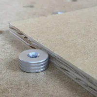Aluminum bench dog with threaded center hole and horizontal grooves, positioned next to a wooden workbench edge with a 20mm hole