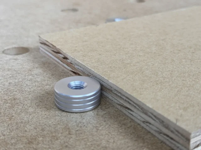 Aluminum bench dog with threaded center hole and horizontal grooves, positioned next to a wooden workbench edge with a 20mm hole