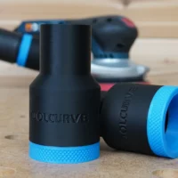 Black ToolCurve hose adapter with blue grip rings shown in pairs, one with tapered end and one with cylindrical end, in focus on wooden surface with Bosch sander blurred in background