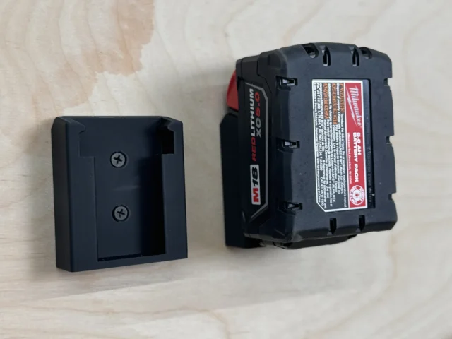 Black plastic battery mount next to a Milwaukee M18 Red Lithium battery, showing the hollow receptacle and mounting holes