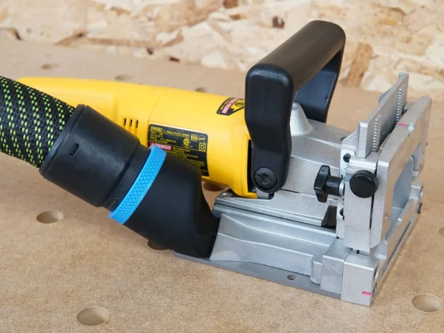 Black hose adapter attached to yellow Dewalt biscuit joiner, mounted on gray fixture with adjustment knobs and metal guides