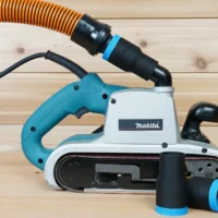 Black ToolCurve 1-1/4 inch hose adapter with blue cuff joining an orange hose to a Makita 9403 belt sander, spare parts in front