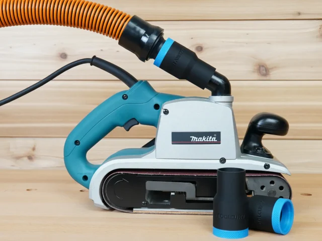 Black ToolCurve 1-1/4 inch hose adapter with blue cuff joining an orange hose to a Makita 9403 belt sander, spare parts in front