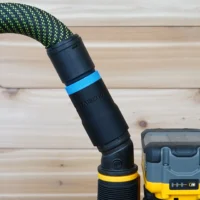 Hose adapter attached to a Dewalt 2-1/4 HP cordless plunge router, showing the black cylindrical connector with blue band and braided green hose above it, next to the yellow and blue router body.