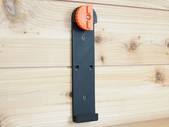Dark gray wall-mounted rail bracket with orange locking clamp at top, mounted vertically on wood with screw holes visible