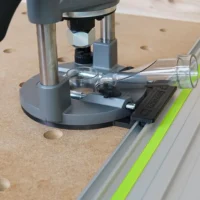 Milwaukee M18 router adapter mounted on Festool guide rail, with clear dust collection tube attached and black clamp visible on the aluminum track