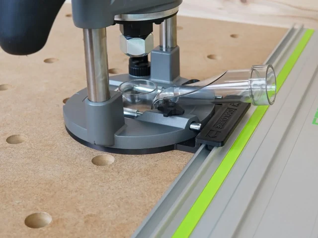 Milwaukee M18 router adapter mounted on Festool guide rail, with clear dust collection tube attached and black clamp visible on the aluminum track